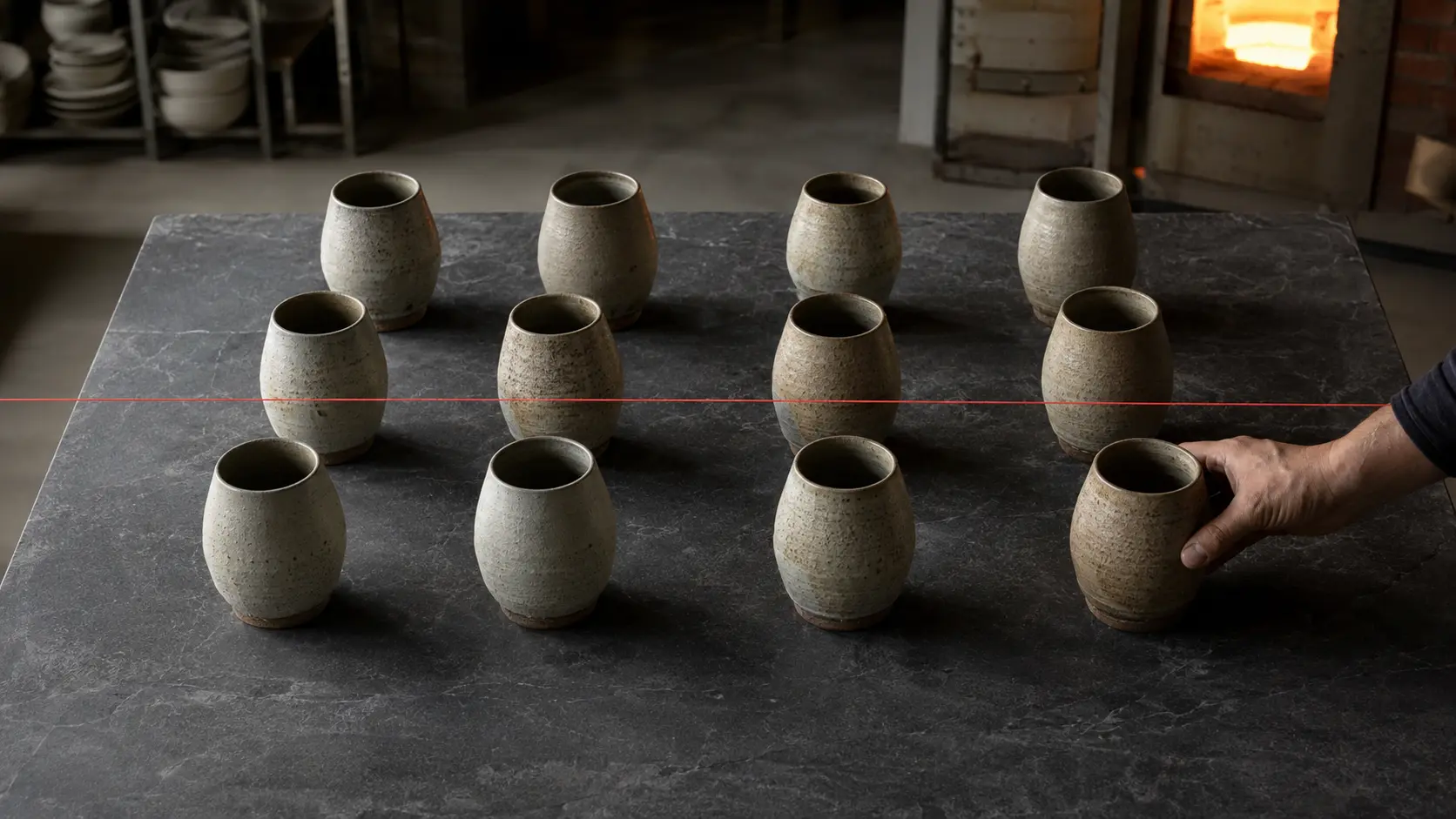 Twelve subtly different handmade stoneware vessels arranged in a measured grid as a hand adjusts the final one
